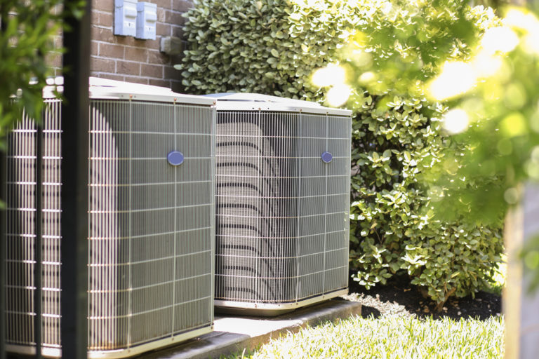 Air conditioner units outside, with tall bushes and a brick house behind them.