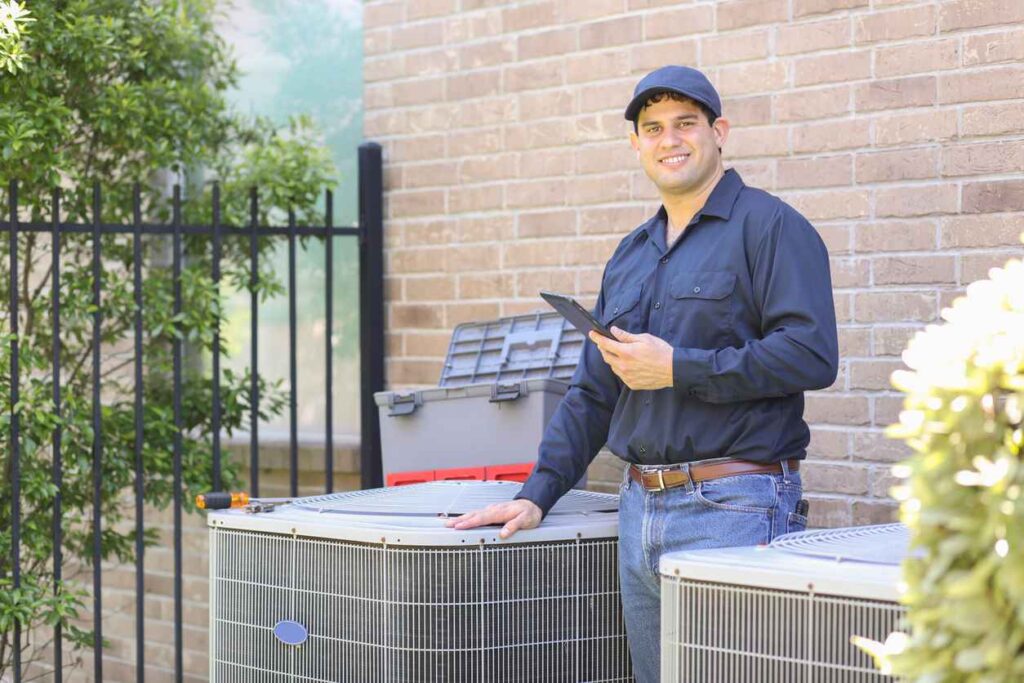 HVAC technician standing behind an air conditioner next to a brick home.