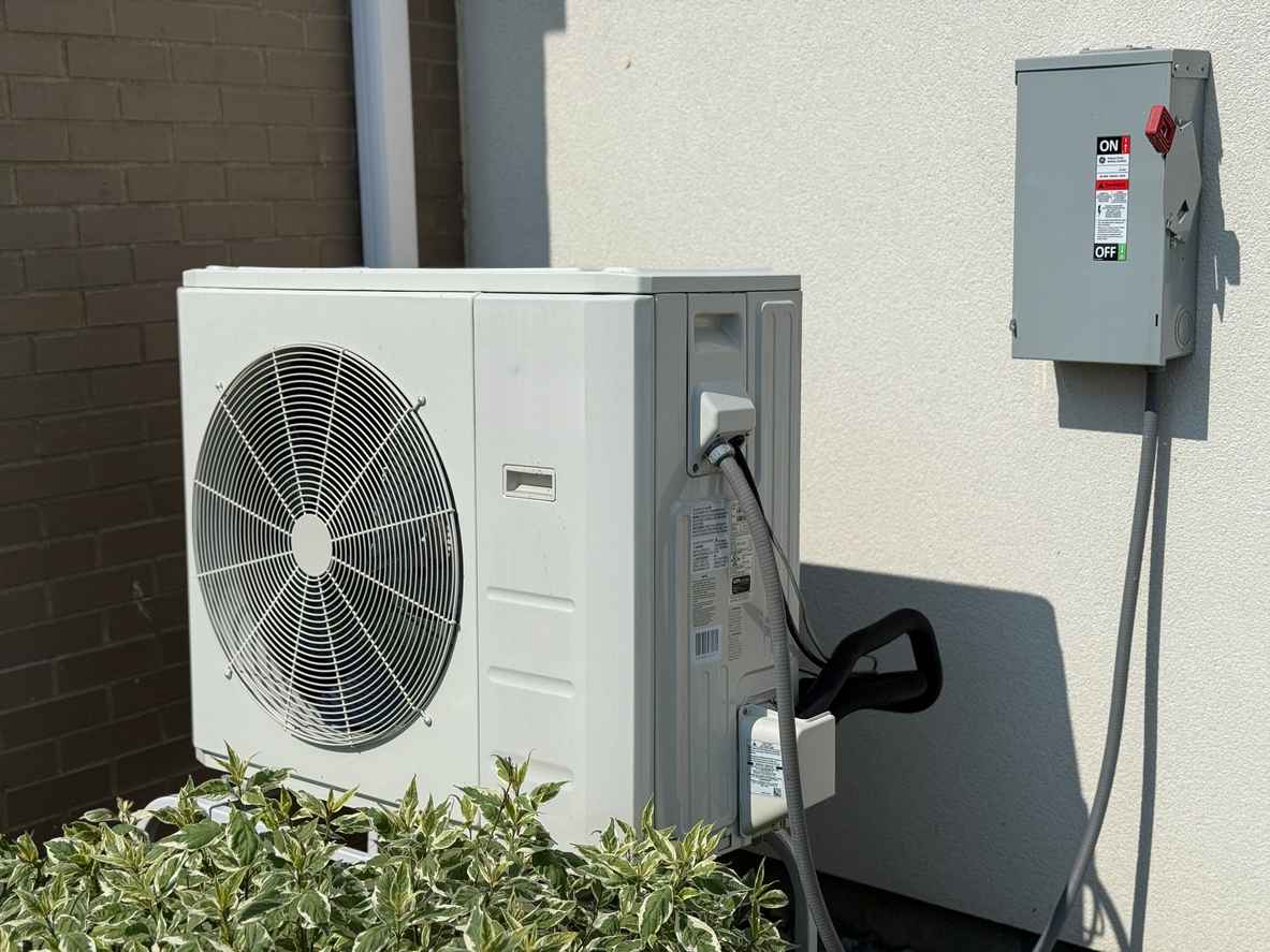 Outdoor HVAC condenser unit beside a stucco wall with a wall-mounted electrical disconnect box nearby.