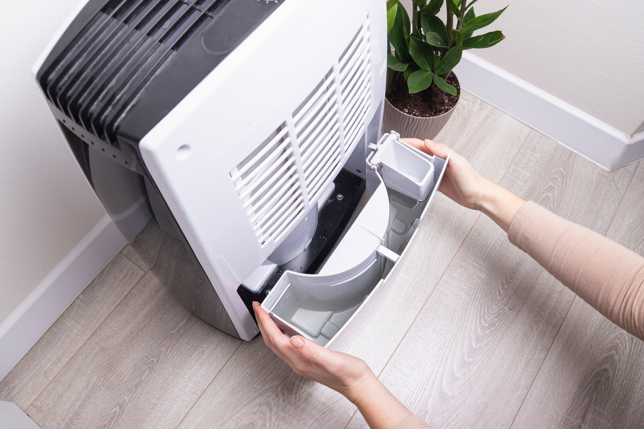 Person removing the water/collection tray from a white portable air conditioner on a wooden floor.